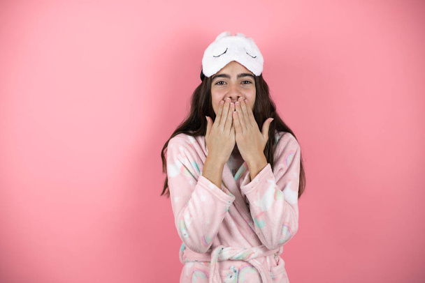 Ładna dziewczyna w piżamie i masce snu na różowym tle zaskoczona rękami nad ustami - Zdjęcie, obraz