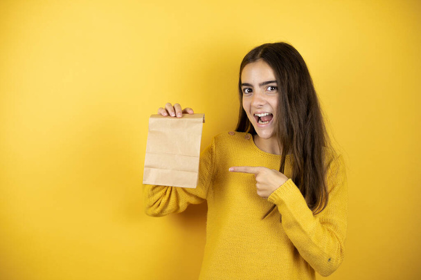 Pretty girl wearing a yellow sweater standing over isolated yellow background holding a paper bag - Photo, Image