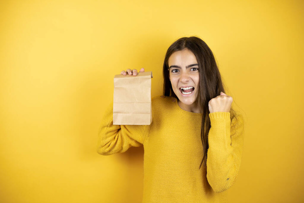 Pretty girl wearing a yellow sweater standing over isolated yellow background holding a paper bag very happy and excited making winner gesture with raised arms, smiling and screaming for success - Foto, afbeelding