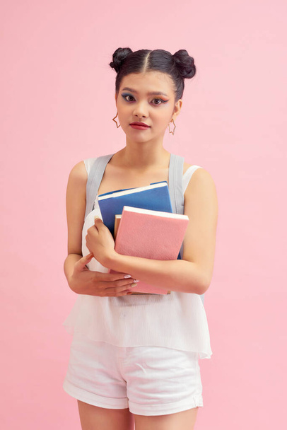 Foto de menina estudante bonita 20s com penteado de pães duplos usando mochila segurando muitos livros de estudo isolados sobre fundo rosa - Foto, Imagem