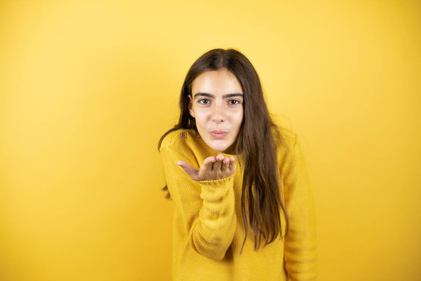Pretty girl wearing a yellow sweater standing over isolated yellow background looking at the camera blowing a kiss with hand on air being lovely and sexy. Love expression. - Fotó, kép