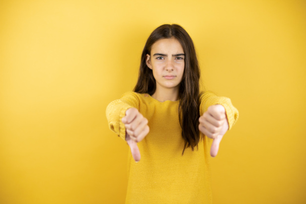 Красивая девушка в желтом свитере стоит на изолированном желтом фоне с сердитым лицом, с негативным знаком неприязни с опущенным большим пальцем - Фото, изображение