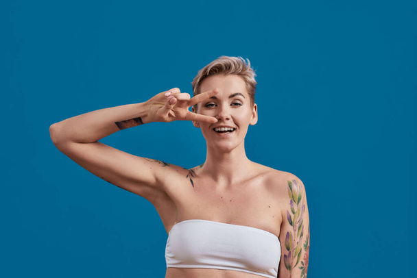 Portrait of a young attractive half naked tattooed woman with perfect skin looking happy, holding hand on eyes and showing victory or peace sign isolated over blue background - Photo, Image