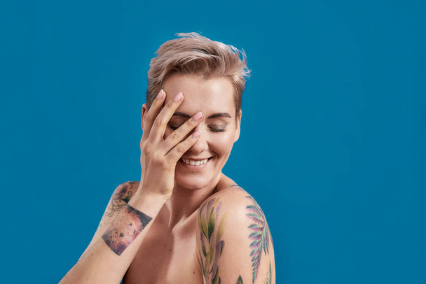 Beauty portrait of a young attractive half naked tattooed woman with perfect skin looking shy with hand over her face isolated over blue background - Fotoğraf, Görsel