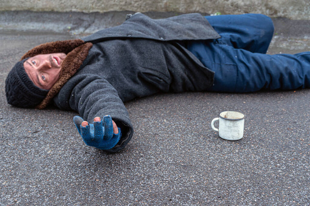Outstretched, dirty hand in a torn blue glove. A homeless man in old dirty clothes lies on the pavement with his hand outstretched. - Photo, Image