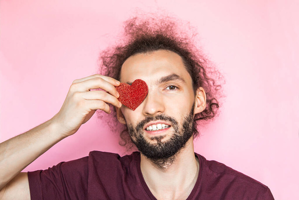 Love and st Valentines day concept. Close-up portrait of smiling handsome bearded man closing eye by red heart, isolated on pink - Fotó, kép