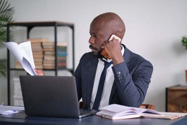 Nuori afrikkalainen johtaja lukee asiakirjoja ja työskentelee kannettavan tietokoneen kanssa istuessaan työpöytänsä ääressä toimistossa. työntekijä tai yrittäjä neuvottelee puhelimitse. pehmeä fokus - Valokuva, kuva