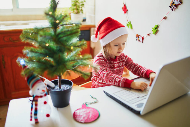 Happy toddler girl in Santa hat with laptop. Kid using computer to communicate with friends or elderly relatives on Christmas or New Year. Celebrating seasonal holidays online while staying at home - Φωτογραφία, εικόνα