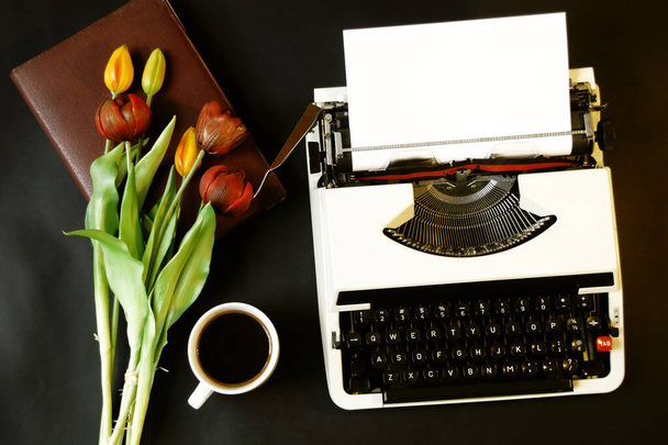 schöne bunte Nahaufnahme von oben Foto von alten Schreibmaschinen, rote und gelbe Tulpen, Tasse Kaffee, rotes Notizbuch auf schwarzem Hintergrund - Foto, Bild