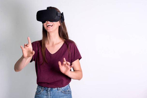 Νεαρή γυναίκα που φοράει γυαλιά εικονικής πραγματικότητας, VR κουτί. Σύνδεση, τεχνολογία, νέα γενιά, έννοια προόδου. Κορίτσι που προσπαθεί να αγγίξει αντικείμενα στην εικονική πραγματικότητα. Στιγμιότυπο στούντιο σε γκρι. - Φωτογραφία, εικόνα