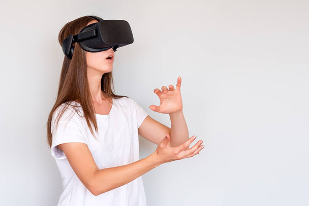 Smiling positive woman wearing virtual reality goggles headset, vr box. Connection, technology, new generation, progress concept. Girl trying to touch objects in virtual reality. Studio shot on gray. - 写真・画像