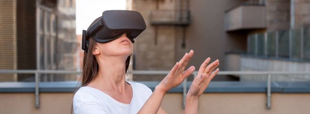 Молода жінка в гарнітурі окулярів віртуальної реальності, коробка vr на відкритому повітрі. Зв'язок, технології, нове покоління, концепція прогресу. Дівчина намагається торкнутися об'єктів у віртуальній реальності
. - Фото, зображення
