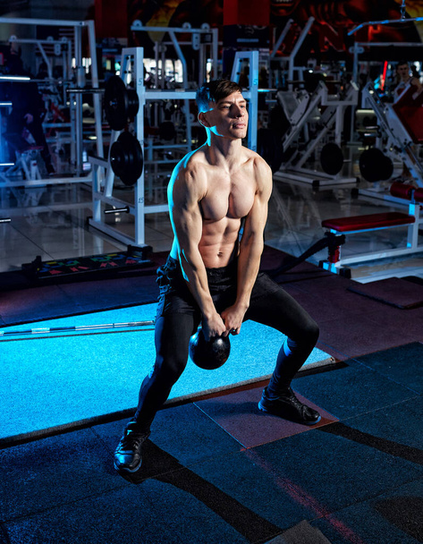 Jeune homme musclé avec torse nu travaillant dans la salle de gym. Athlétisme masculin adulte faisant de l'exercice avec cloche de bouilloire. Fitness, concept sportif. Rétro-éclairage bleu - Photo, image