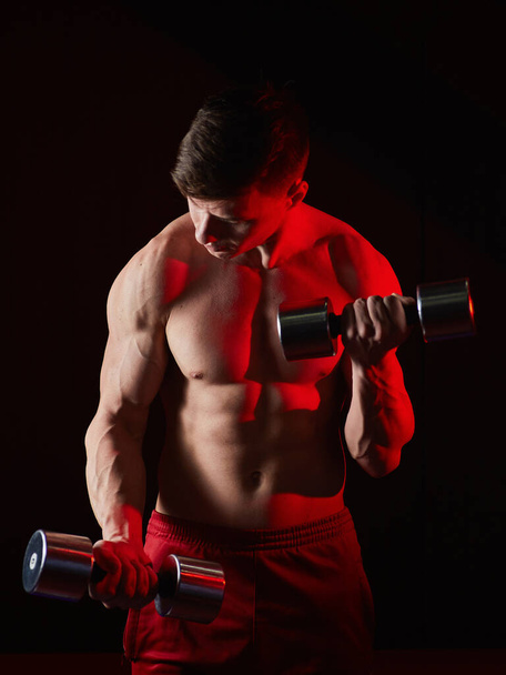 Sporty young man training with dumbbells against dark background. Red background light - Foto, Imagen