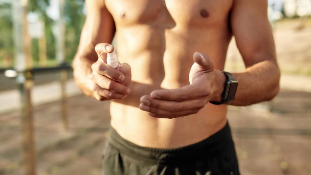 Close up of muscular young half naked guy using sanitizer on hands while having workout at outdoor gym. Person protecting himself against coronavirus - Foto, afbeelding