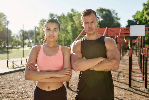 Portrait of muscular young guy and sportive girl looking at camera, standing with arms crossed while exercising together at outdoor gym - Foto, afbeelding