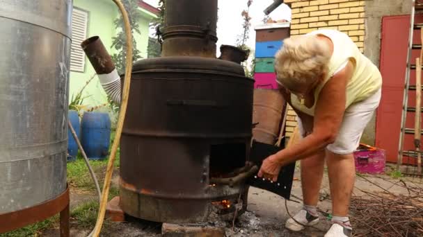 Grandma throws dry branches into the firebox of a homemade distillery making moonshine schnapps, alcoholic beverages. - Footage, Video