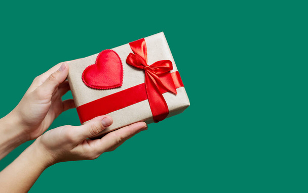 női kéz gazdaság barna ajándék doboz piros íj és a szív a háttérben a zöld szín.másolás tér - Fotó, kép