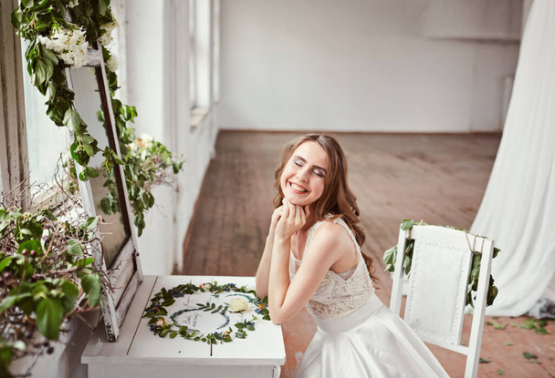 Piękna panna młoda w białej sukience siedzi na krześle przy oknie i trzyma bukiet ślubny. Białe studio. - Zdjęcie, obraz