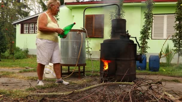 おばあちゃんは、蒸留装置の下で火を鎮めるために、国内のアルコール飲料を作るために、熱を減らすために水のボトルで. - 映像、動画
