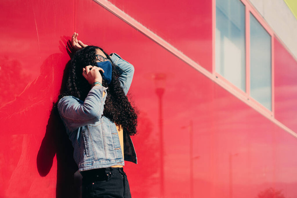 LGTB transznemű lány göndör hajjal és arcmaszkkal telefonál egy piros falon állva. A koronavírus fogalma. - Fotó, kép