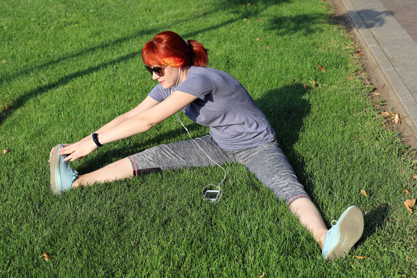 Jeune femme aux cheveux roux en tenue de sport s'assoit sur la pelouse et effectue des exercices d'étirement tout en écoutant de la musique sur écouteurs le jour ensoleillé - Photo, image
