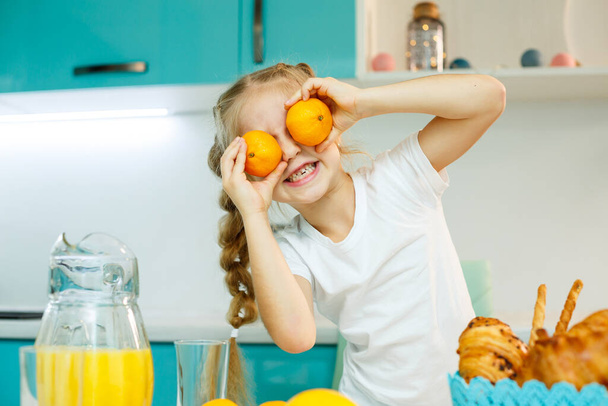A girl of 7-8 years old sits in the kitchen and makes eyes with a tangerine. - Fotoğraf, Görsel
