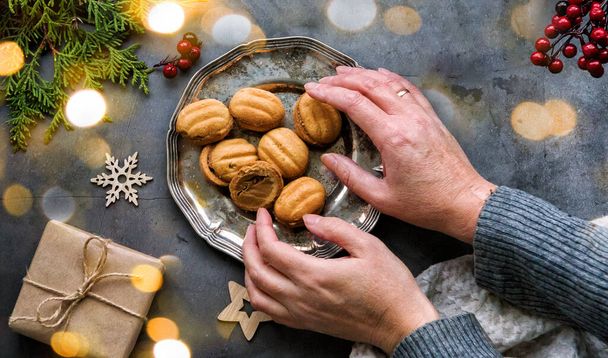 Las manos elegantes de las mujeres tocan un plato de galletas-nueces en una mesa oscura. Diseño enmarcado de decoraciones navideñas, regalos, nueces y árbol de thuja. Diseño de Navidad - Foto, Imagen