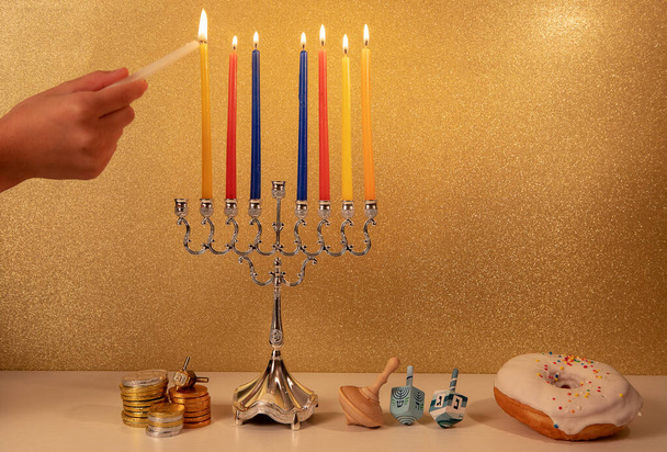 Día 7 de fiesta religiosa judía Hanukkah con la mano del niño encendiendo la séptima vela en la tradicional menorá araña sobre fondo dorado brillante con otros objetos tradicionales alrededor... - Foto, imagen