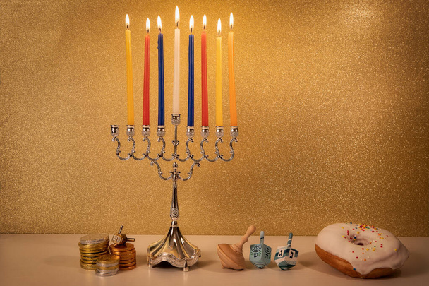 Day 7 of jewish religious holiday Hanukkah with traditional chandelier menorah, spinning top toys (dreidels) and a doughnut and chocolate coins on white wooden table and golden glittering background - Photo, Image