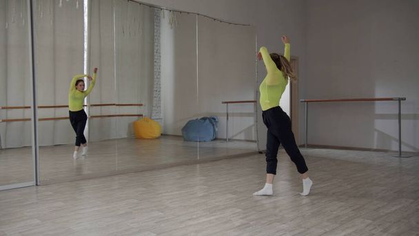 Ένα όμορφο κορίτσι χορεύει μπροστά από έναν καθρέφτη και preens κατά τη διάρκεια μιας άσκησης χορού στο γυμναστήριο - Φωτογραφία, εικόνα