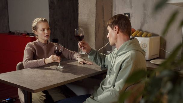 Ένα νεαρό παντρεμένο ζευγάρι πίνει κόκκινο κρασί στην κουζίνα του, φτιάχνει μια πρόποση και βάζει ποτήρια. - Φωτογραφία, εικόνα