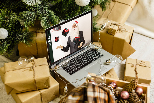 Ordenador en la mesa en la acogedora habitación con Santa Claus en la pantalla que desea Feliz Navidad y Feliz Año Nuevo en línea y haciendo milagro hecho realidad - Foto, Imagen