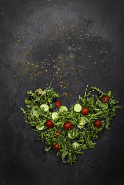 Fresh vegetables arugula, tomato, cucumber, greens, peppercorns and olives on a dark gray textured background in the form of a heart. View from above. Place for text, copy space. - Foto, Imagem