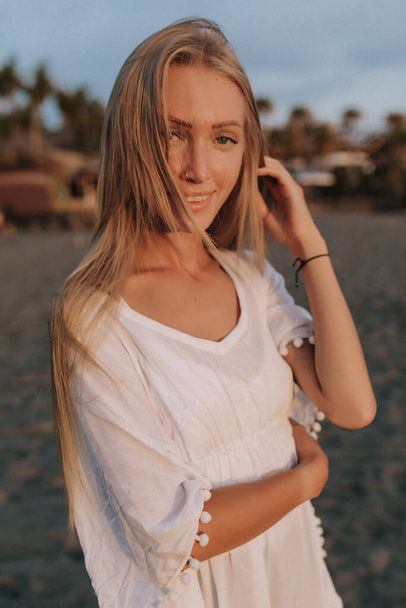 Csinos hölgy hosszú szőke hajjal, fehér pólóban, aki az óceán partján sétál napfényben. - Fotó, kép