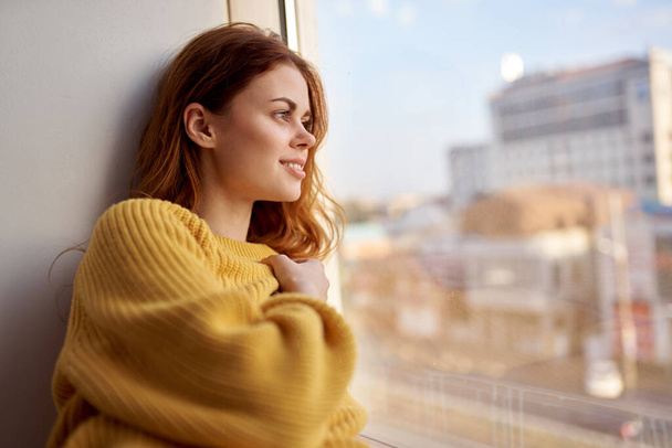 una mujer bonita en un suéter amarillo se encuentra en el alféizar de la ventana y mira por la ventana - Foto, Imagen