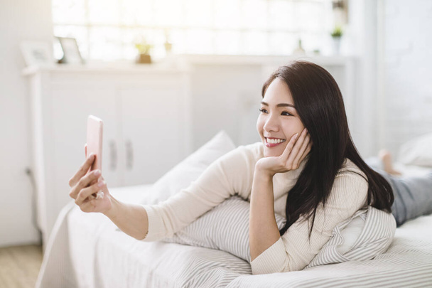 Щаслива молода жінка, використовуючи відеодзвінок по мобільному телефону і лежачи на ліжку
  - Фото, зображення