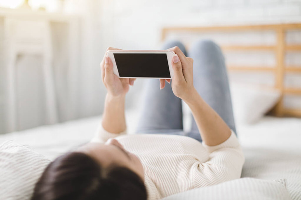 Nuori nainen katsomassa matkapuhelimen näytön ja makaa sängyllä kotona - Valokuva, kuva