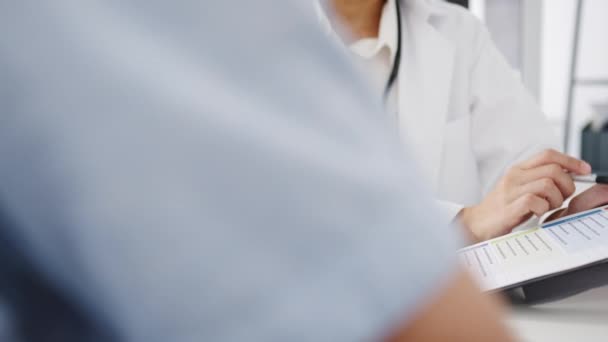 Junge asiatische Ärztin in weißer medizinischer Uniform mit Klemmbrett liefert großartige Nachrichten sprechen über Ergebnisse oder Symptome mit männlichen Patienten, die am Schreibtisch in der Klinik oder im Krankenhaus sitzen. - Filmmaterial, Video