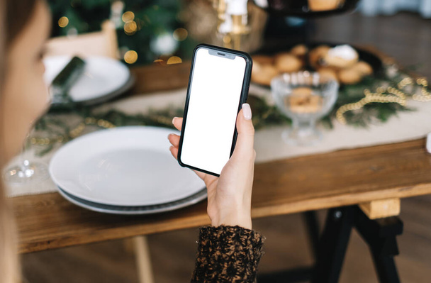 Woman holding smartphone with a white screen mock up at home near festive table - Foto, afbeelding