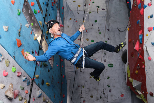 Профессиональный старший человек летит после прыжка, пока он взбирается на искусственную стену скалолазания. Концепция экстремального спорта. - Фото, изображение