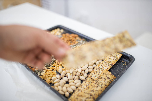 Female hand holding multi-seed crispbread above a tray with seeds and nuts - Фото, изображение