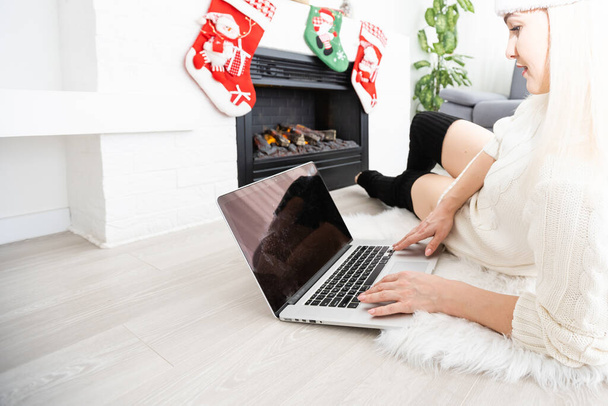 Mujer joven y feliz navegando por Internet durante la temporada navideña mientras se sienta en el suelo en la sala de estar frente a una Navidad decorada - Foto, imagen