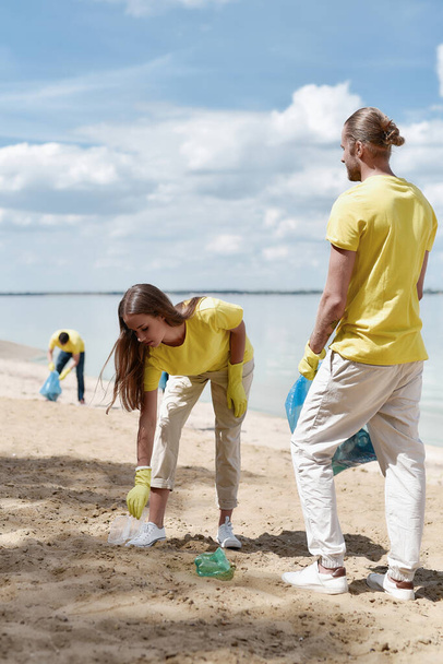 Two young eco activists wearing uniform and rubber gloves collecting plastic waste on the beach with group of volunteers - Foto, afbeelding