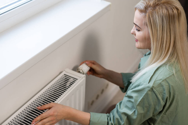 Young and beautiful blond woman touching the radiator. Heating season concept. - Photo, Image
