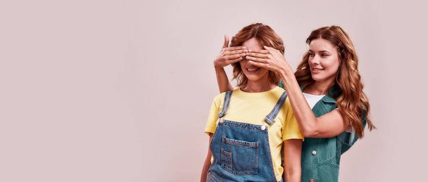 Portrait of two attractive twin sisters in casual wear. Young girl covering sisters eyes with hands to surprise her, standing isolated over light background - Foto, afbeelding