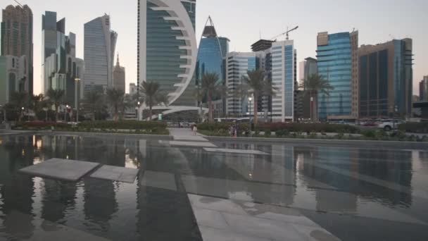 Westelijke baai wolkenkrabbers in Doha Qatar middag panning schot genomen uit Sheraton park met fontein op de voorgrond, Qatar vlaggen, palmbomen en auto 's bewegen in de Corniche straat - Video
