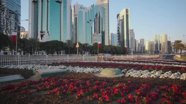 Zachodnia zatoka drapacze chmur w Doha Katar popołudniowe powiększenie zdjęć zrobione z parku Sheraton z kwiatami, flagi Kataru na pierwszym planie i samochody poruszające się na ulicy Corniche - Materiał filmowy, wideo