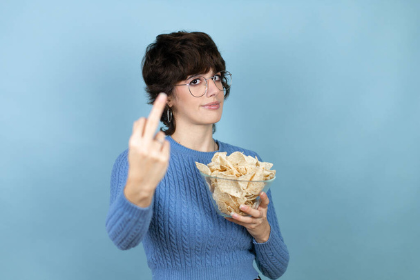 Young beautiful woman holding nachos potato chips over isolated blue background showing middle finger doing fuck you bad expression, provocation and rude attitude. screaming excited - Photo, Image