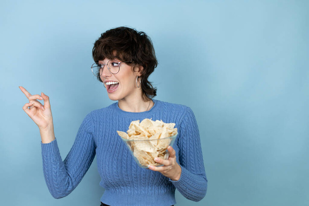 Young beautiful woman holding nachos potato chips over isolated blue background very happy pointing with hand and finger to the side - Foto, Imagem